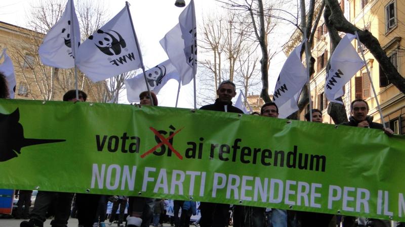 Un gruppo di persone tiene uno striscione con la scritta “Vota sì al referendum”. Alcuni partecipanti sventolano bandiere del WWF, caratterizzate dal logo del panda stilizzato bianco e nero.